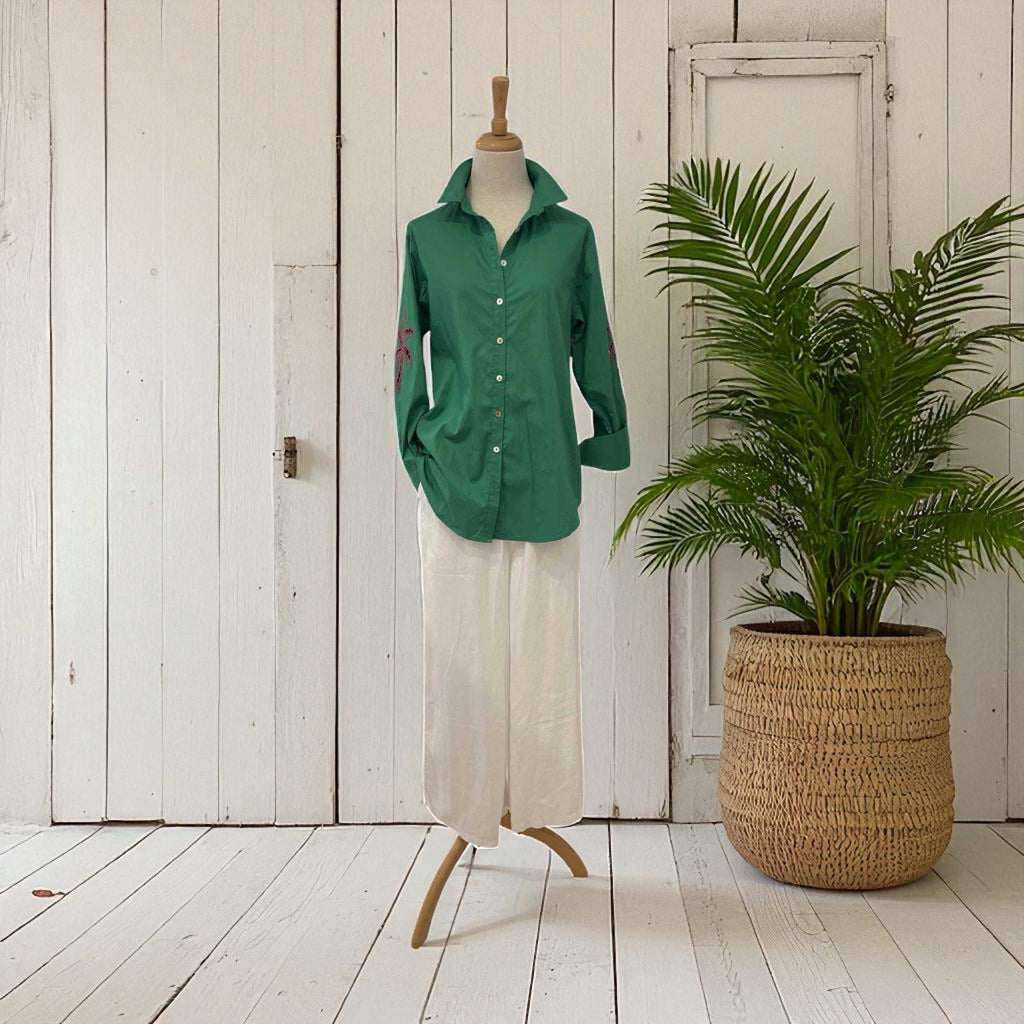 Green shirt on a mannequin with beige pants, next to a potted plant against a white wooden wall.