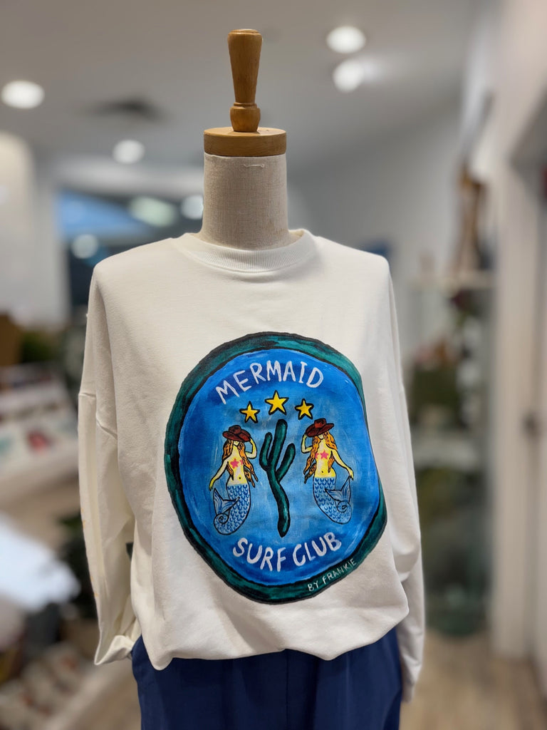 White sweatshirt with a 'Mermaid Surf Club' patch on a mannequin in a store setting.