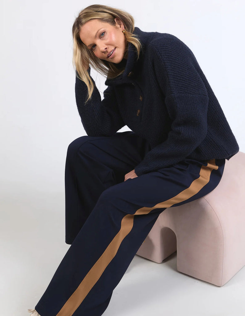 Person wearing a navy tracksuit with a brown stripe sitting on a pink stool against a white background